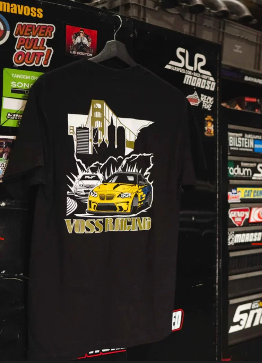 Back of Voss Racing Tee Black with a yellow racing car graphic and 'Voss Racing' text, displayed against a backdrop of automotive-themed posters.